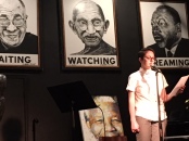 Tanya in white button-down shirt in front of a microphone holding a piece of paper, reading a poem on a black stage, portraits of Dalai Lama, Gandhi, and MLK behind her